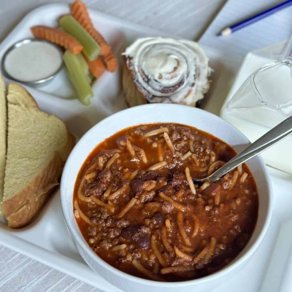 A school tray with chili, veggies, a cinnamon roll, sandwich and milk.