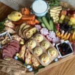 This is a picture of a charcuterie board. On it there are crackers, cheddar cheeses, deviled eggs, a rainbow colored fruit assortment, raw veggies, a ranch dip, mixed nuts, dried fruit and pimento cheese stuffed carrots.