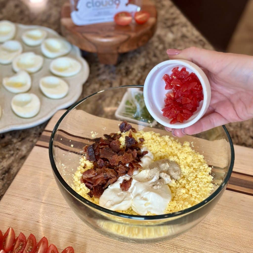 Pouring diced tomato into a bowl with mashed egg yolk and other ingredients