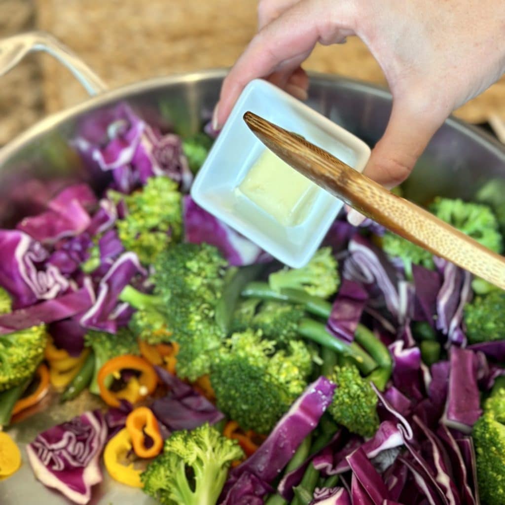 Adding butter to a skillet of vegetables.