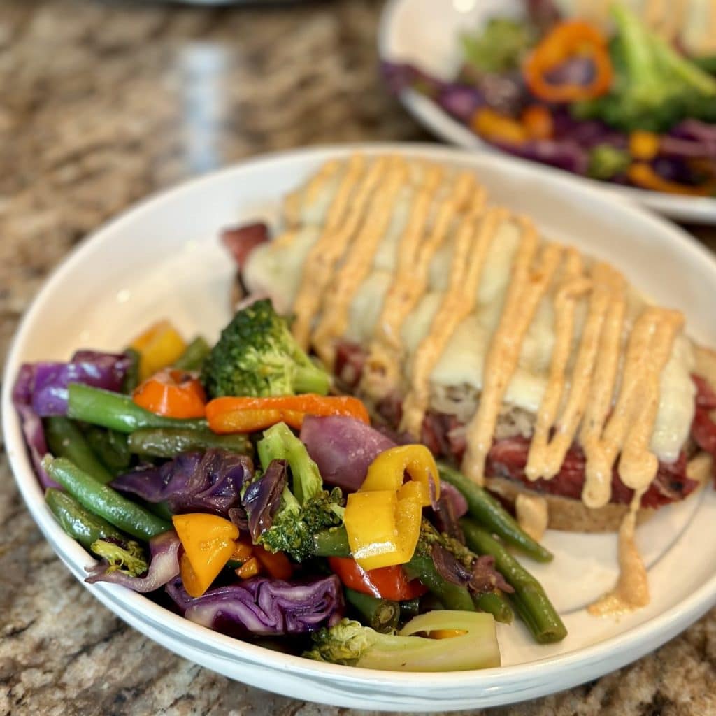 A dinner plate topped with an open face pastrami sandwich and cooked vegetables.