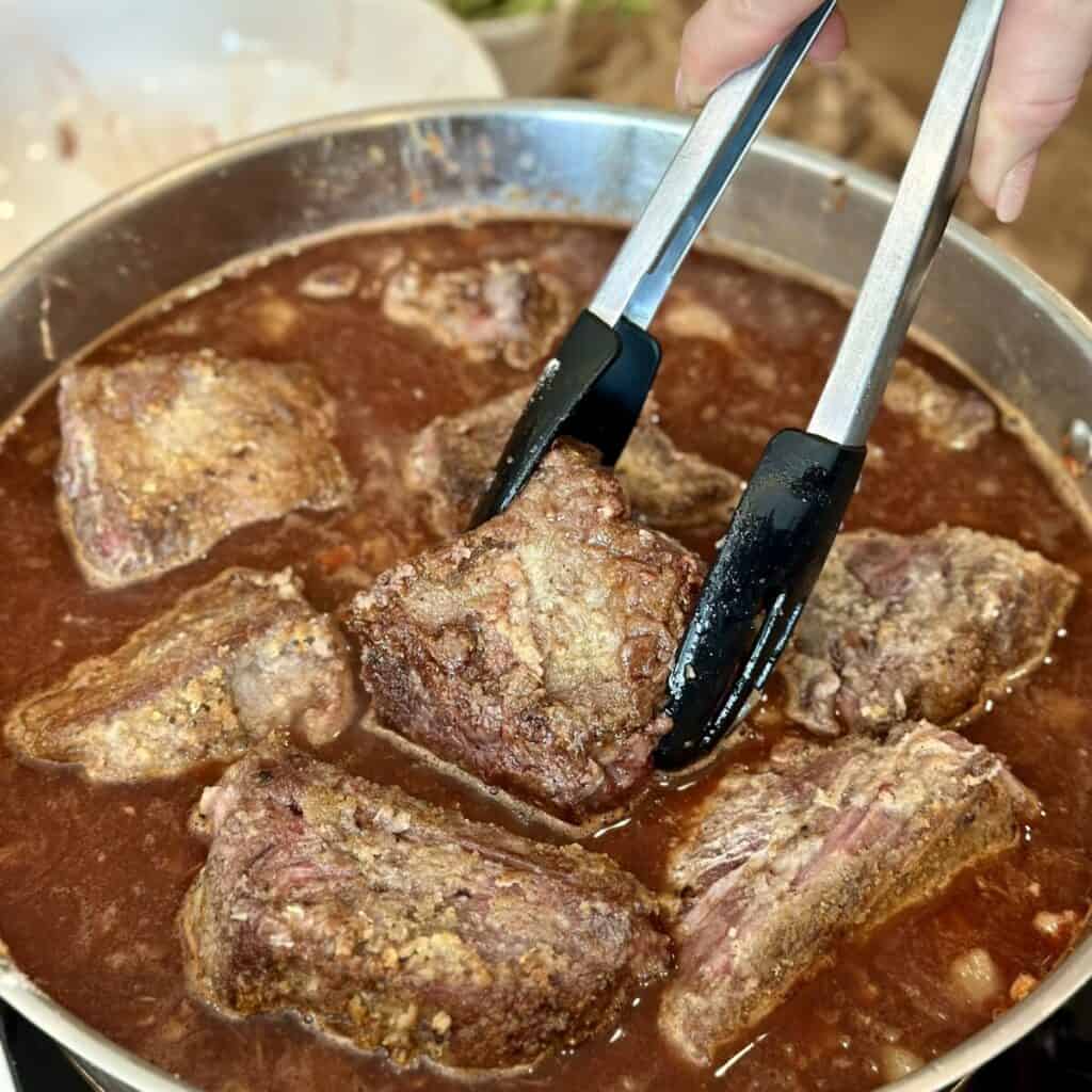 Adding seared short ribs to a pan of broth and veggies.