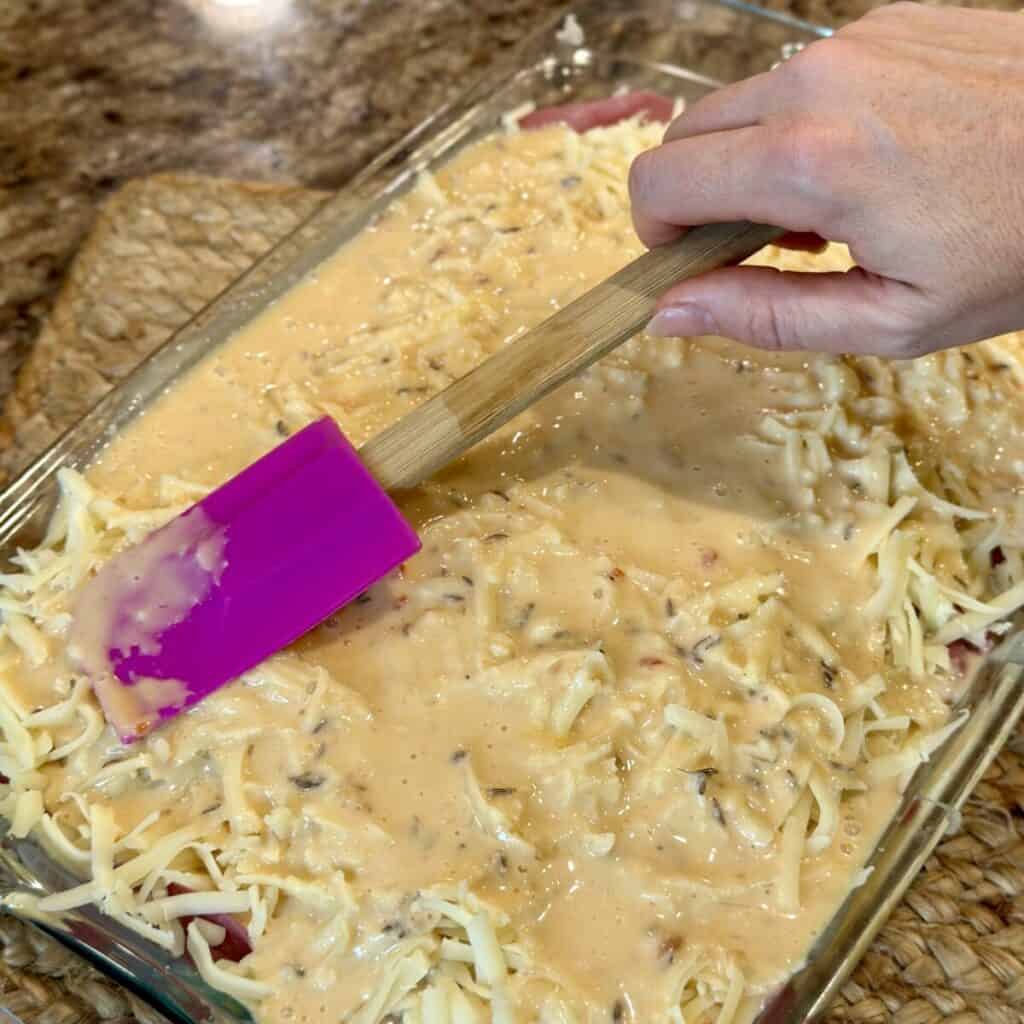 Adding the liquid mixture to the top of a Reuben casserole.