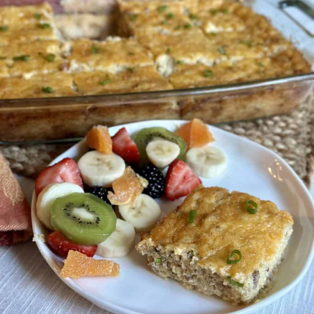 A slice of grits and sausage casserole on a plate with fresh fruit.