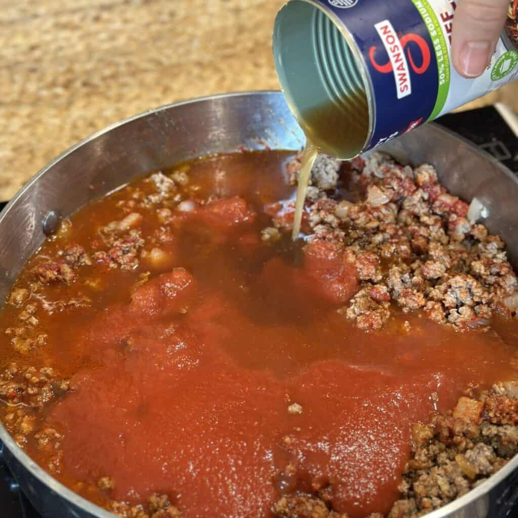 Adding broth to beef and tomato sauce in a skillet.